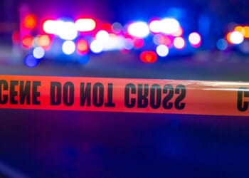 Police lights flash behind tape at a crime scene at night.