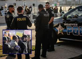 A group of local police officers watch from across the street as demonstrators gather and sing in the "free speech zone" outside the Immigration and Customs Enforcement processing and detention facility on October 31, 2025, in Broadview, Illinois. The Village of Broadview established the restricted protest zone and set curfew hours amid President Donald Trump