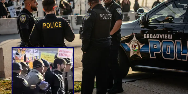 A group of local police officers watch from across the street as demonstrators gather and sing in the "free speech zone" outside the Immigration and Customs Enforcement processing and detention facility on October 31, 2025, in Broadview, Illinois. The Village of Broadview established the restricted protest zone and set curfew hours amid President Donald Trump