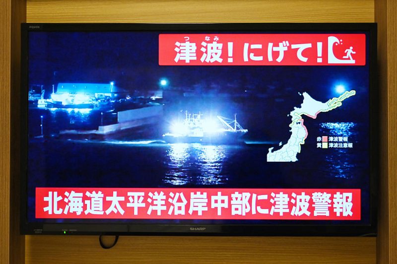 TOPSHOT - An advisory reading "Tsunami! Run!" (top) and "Tsunami warning issued for central Hokkaido's Pacific Coast" flashes over the live footage of a boat leaving a port, on a television screen in Sapporo, in Japan's northern Hokkaido prefecture on December 8, 2025. Japan recorded a 40-centimetre (16-inch) tsunami after a 7.6-magnitude quake on December 8. (Photo by GREG BAKER / AFP via Getty Images)