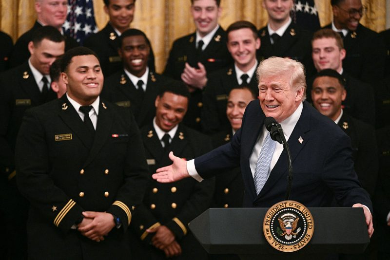 US President Donald Trump gestures to Navy defensive tackle Landon Robinson (L) while speaking during a ceremony to present the Commander-in-Chief Trophy to the Navy Midshipmen football team of the United States Naval Academy in the East Room of the White House in Washington, DC, on March 20, 2026. (Photo by Brendan SMIALOWSKI / AFP via Getty Images)