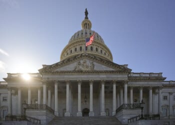 U.S. Capitol with sun behind the dome and an American flag flying against a clear blue sky.