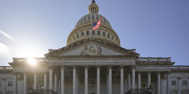 U.S. Capitol with sun behind the dome and an American flag flying against a clear blue sky.