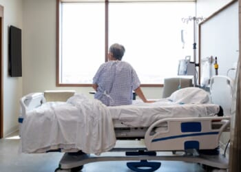 A photo illustration depicts an older woman seated on a hospital bed alone in her room.